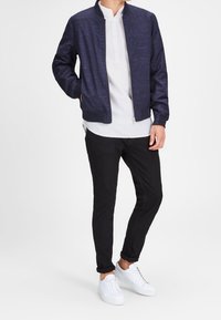 Giacca bombardiere blu navy con tessuto a trama, polo bianca, pantaloni neri a sigaretta e sneakers bianche. La giacca presenta un collo rialzato e tasche laterali.