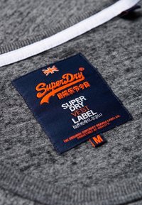 Etiqueta de chaleco SuperDry en azul marino cosida sobre un tejido gris texturizado, con texto en naranja y blanco y un pequeño emblema de bandera en naranja y blanco.