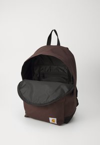 Brown backpack made of durable fabric, featuring a black flap, top handle, adjustable straps, and an interior zip pocket.