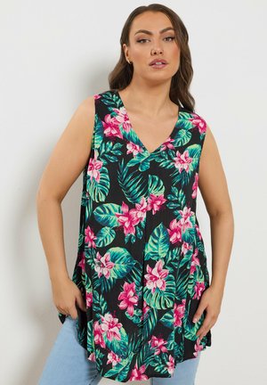 Frau trägt ein ärmelloses schwarzes Oberteil mit pink-grünem Blumenmuster, steht vor einem schlichten weißen Hintergrund.