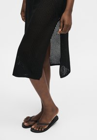Personne portant une jupe midi noire en tricot avec une fente latérale et des tongs noires, debout sur fond blanc.