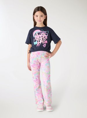 Ragazza giovane in piedi con la mano sul fianco, indossa una maglietta blu navy con scritta "Good Day", pantaloni tie-dye multicolore pastello e scarpe da ginnastica rosa-bianco.
