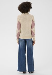 Ženska s kodrasto frizuro, oblečena v bež pulover z rožnatimi diamanti na rokavih in širokimi modrimi kavbojkami, stoji s hrbtom obrnjena.