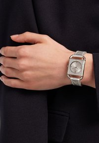 Sølv rektangulært ur med en tekstureret læderrem, der har en enkel hvid skive og minimalistiske timemarkører, båret på en hånd.