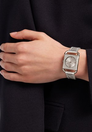 Sølv rektangulært ur med en tekstureret læderrem, der har en enkel hvid skive og minimalistiske timemarkører, båret på en hånd.