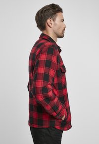 Camisa de franela a cuadros rojos y negros con mangas largas, cierre de botones en la parte delantera y un bolsillo en el pecho, hecha de una tela texturizada.