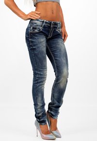 Jeans skinny de mezclilla azul con áreas desgastadas, costuras de contraste y bolsillos frontales, combinados con tacones altos brillantes de múltiples colores.