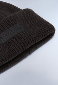 Beanie nera in maglia con un motivo testurizzato. Presenta una toppa nera rettangolare sulla parte anteriore. Materiale morbido e flessibile.