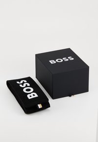 Crna kutija s bijelim logotipom "BOSS", koja sadrži crnu suknu torbicu s logotipom, zlatnim i bijelim naglascima. Kutija je kvadratnog oblika.