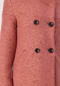 Casaco texturizado rosa com design de dupla matelassado, botões pretos e um bolso lateral. O tecido parece quente e ligeiramente felpudo.