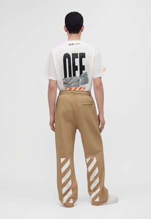 Personne portant un T-shirt blanc avec le texte et le graphisme "OFF" dans le dos, associé à un pantalon beige avec des rayures blanches diagonales sur les jambes.