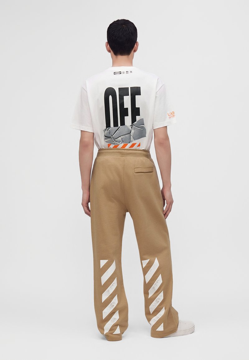 Άτομο φοράει λευκό T-shirt με κείμενο και γραφικό "OFF" στην πλάτη, σε συνδυασμό με μπεζ παντελόνια με διαγώνιες λευκές ρίγες στα πόδια.