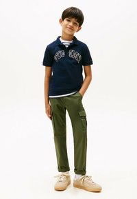 Marine polo med "TOMMY" branding, grøn cargo bukser med sidelommer, lyse sneakers, stribet skjorte synlig ved kraven.