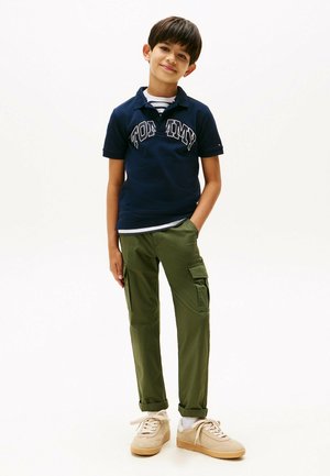 Navy polo with "TOMMY" branding, green cargo pants with side pockets, light sneakers, striped inner shirt visible at the collar.