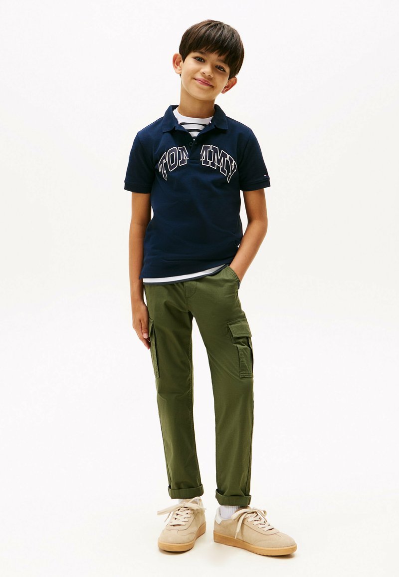 Marine polo med "TOMMY" branding, grøn cargo bukser med sidelommer, lyse sneakers, stribet skjorte synlig ved kraven.