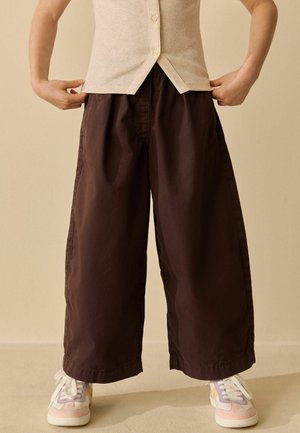 Person wearing wide-leg dark brown pants, beige ribbed buttoned top, and pastel-colored sneakers, standing against a plain background.