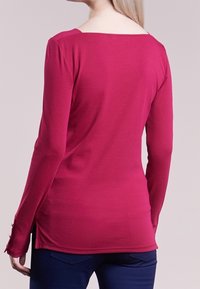 Långärmad, fuchsia topp med båt-uttäckning, sidofickor och slät yta. Bakifrån visas en figursydd form och sömmar.