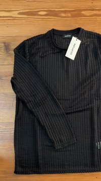 Top preto de mangas longas com um padrão texturizado em forma de grelha e decote redondo. Inclui uma etiqueta presa ao colarinho.
