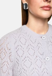 Helle lavendelfarbene Strickjacke mit einem Rautenmuster, das eine strukturierte Oberfläche aufweist und drei weiße Knöpfe am Halskragen hat.