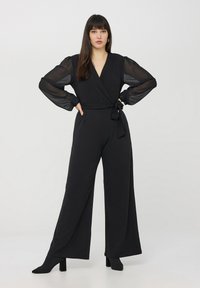Czarny kombinezon z wiązanym fasonem, z przezroczystymi długimi rękawami i wiązaniem w talii. Materiał ma gładką teksturę, uzupełniony o botki na obcasie.