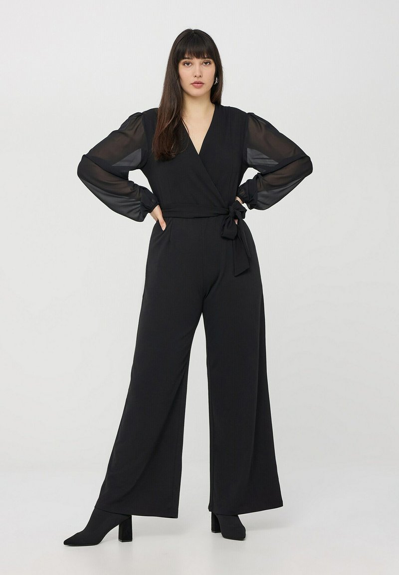 Czarny kombinezon z wiązanym fasonem, z przezroczystymi długimi rękawami i wiązaniem w talii. Materiał ma gładką teksturę, uzupełniony o botki na obcasie.