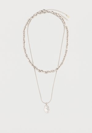 Deux colliers en argent, l'un à chaîne épaisse et l'autre à chaîne fine avec un pendentif de forme irrégulière, posés à plat sur un fond blanc.