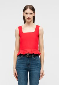 Mujer con una camiseta sin mangas roja acanalada con dobles volantes y vaqueros azules de talle alto con un cinturón negro, sobre un fondo liso.