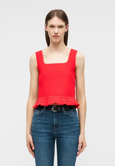 Γυναίκα που φοράει κόκκινο αμάνικο πλεκτό τοπ με βολάν στο τελείωμα και τζιν ψηλόμεσο μπλε με μαύρη ζώνη, μπροστά σε απλό φόντο.