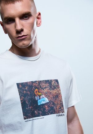 Giovane uomo con capelli corti che indossa una maglietta bianca con la foto di una tomba con fiori e la parola "FORMER" in basso a destra.