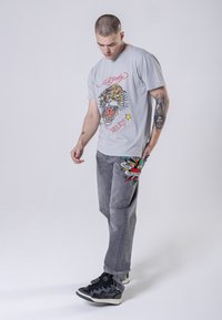 Šedé grafické tričko s barevným designem tygra, spárované se šedými džínami zdobenými barevným orlem a textovými grafikami. Černé tenisky.