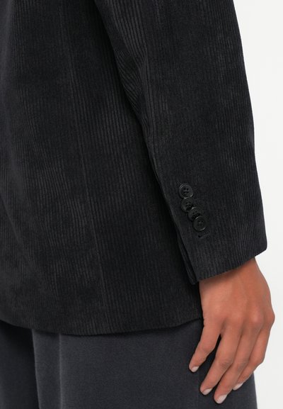 Veste en velours côtelé noir avec un motif de côtes verticales texturé. Dispose d'un poignet à boutons avec quatre boutons noirs et d'une coupe droite.