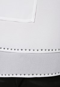 Top bianco con una texture liscia, caratterizzato da un bordo orizzontale decorato con borchie metalliche lungo il bordo inferiore.