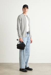 Cardigan grigio a coste, t-shirt bianca, jeans a gamba larga blu chiaro, scarpe nere, con una piccola borsa a tracolla nera, il tutto su uno sfondo neutro.