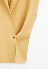 Hellbeige einreihiger Blazerärmel mit Knopf und Vorderansicht vor einem schlichten weißen Hintergrund.