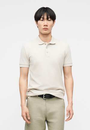 Tânăr cu păr negru drept, purtând un tricou polo bej cu mâneci scurte, băgat în pantaloni kaki deschiși, cu o curea neagră.