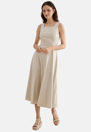 Mujer con vestido midi beige sin mangas con escote cuadrado y sandalias de cuña beige, de pie con las manos ligeramente entrelazadas.