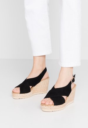 Pieds portant des sandales à talons compensés en cuir de velours noir avec des bretelles croisées et une boucle à la cheville, associés à un pantalon court blanc sur un fond clair.