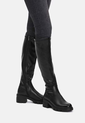 Czarne skórzane kozaki do kolan z bocznym zamkiem, noszone z ciemnymi obcisłymi dżinsami, ujęte pod kątem, aby pokazać zarówno wewnętrzne, jak i zewnętrzne strony.