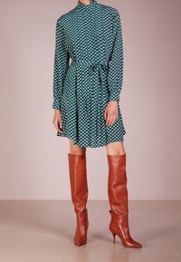 Långärmad grön klänning med vitt geometriskt mönster, åtsittande midja och knälång kjol, i kombination med glansiga bruna stövlar som går upp till låren.