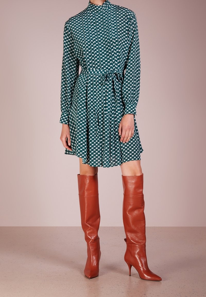 Långärmad grön klänning med vitt geometriskt mönster, åtsittande midja och knälång kjol, i kombination med glansiga bruna stövlar som går upp till låren.