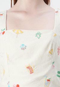 Rochie brodată cu decolteu pătrat, dintr-un material bej deschis, cu modele colorate inspirate din vară, inclusiv scoici și băuturi.