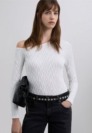 Witte top met lange mouwen van gestructureerde stof, off-shoulder ontwerp, gecombineerd met een zwarte, hooggetailleerde jeans en een zwarte leren clutch.
