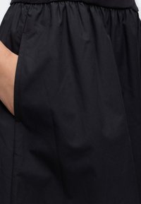 Χέρι που στηρίζεται στην αριστερή τσέπη ενός φαρδιού μαύρου παντελονιού με ελαστική ζώνη μέσης.