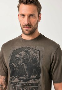 Hombre con cabello corto y barba que lleva una camiseta marrón con la imagen de un oso rugiente atrapando un pez y el texto "Glacier Bay Alaska".
