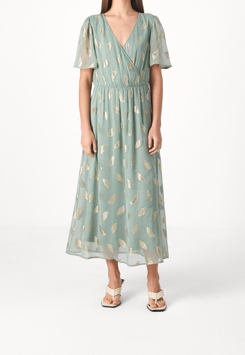 Femme portant une robe midi vert clair avec des motifs de feuilles dorées, des manches flottantes, un décolleté en V et des sandales à talons beiges.