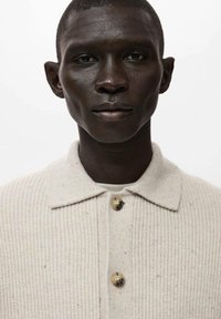 Beżowy, prążkowany sweter z kołnierzykiem i zapięciem na guziki. Charakteryzuje się teksturowaną tkaniną i jasnymi włóknistymi plamkami.