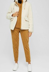 Kreemi fleece takki hupun kanssa, vetoketjukiinnitys ja tuplataskut edessä, yhdistettynä ruskeaan ribbipaitaan ja sinapinkeltaisiin samettihousuihin. Valkoiset tennarit.