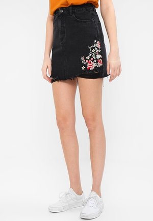 Sort denim miniskørt med frynset kant og blomsterbroderi i pink og hvid på siden. Båret med en sennepsgul top og hvide sneakers.