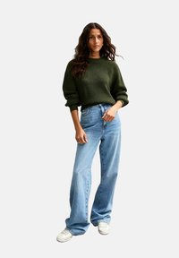 Groene gebreide trui met een relaxte pasvorm, gecombineerd met lichtblauwe wijduitlopende jeans en witte sneakers; zachte textuur en casual ontwerp.