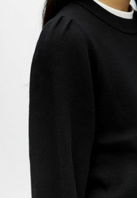 Pull en maille noire avec un col rond et un léger détail bouffant à l'épaule, présentant une texture douce et une silhouette ajustée.